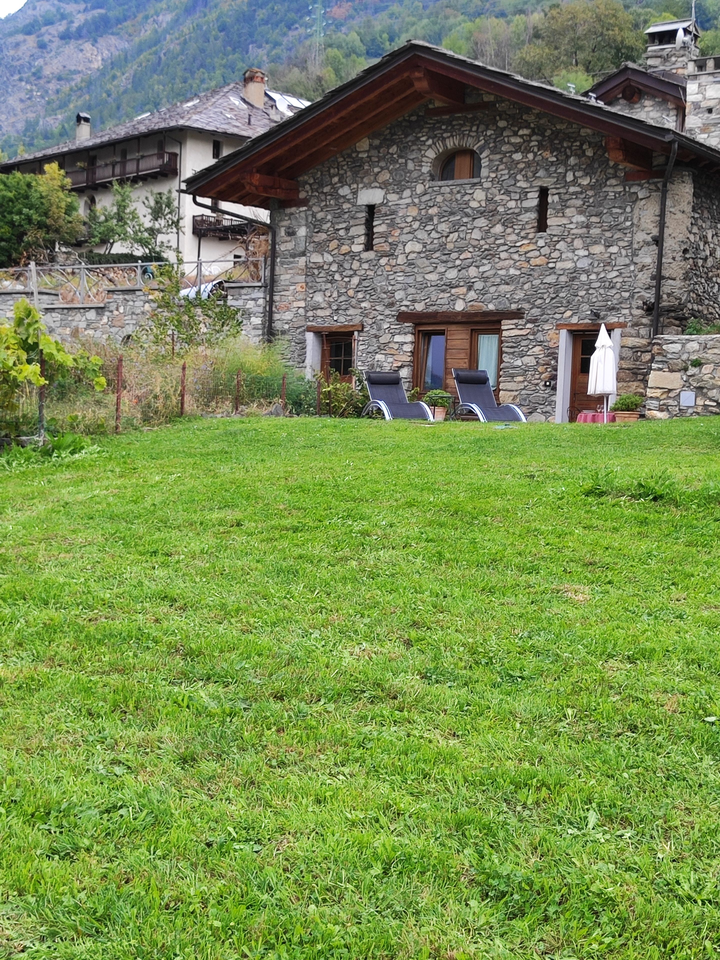 Une maison en pierre avec un toit en pente, un petit porche et deux fauteuils de jardin avec des parasols.
