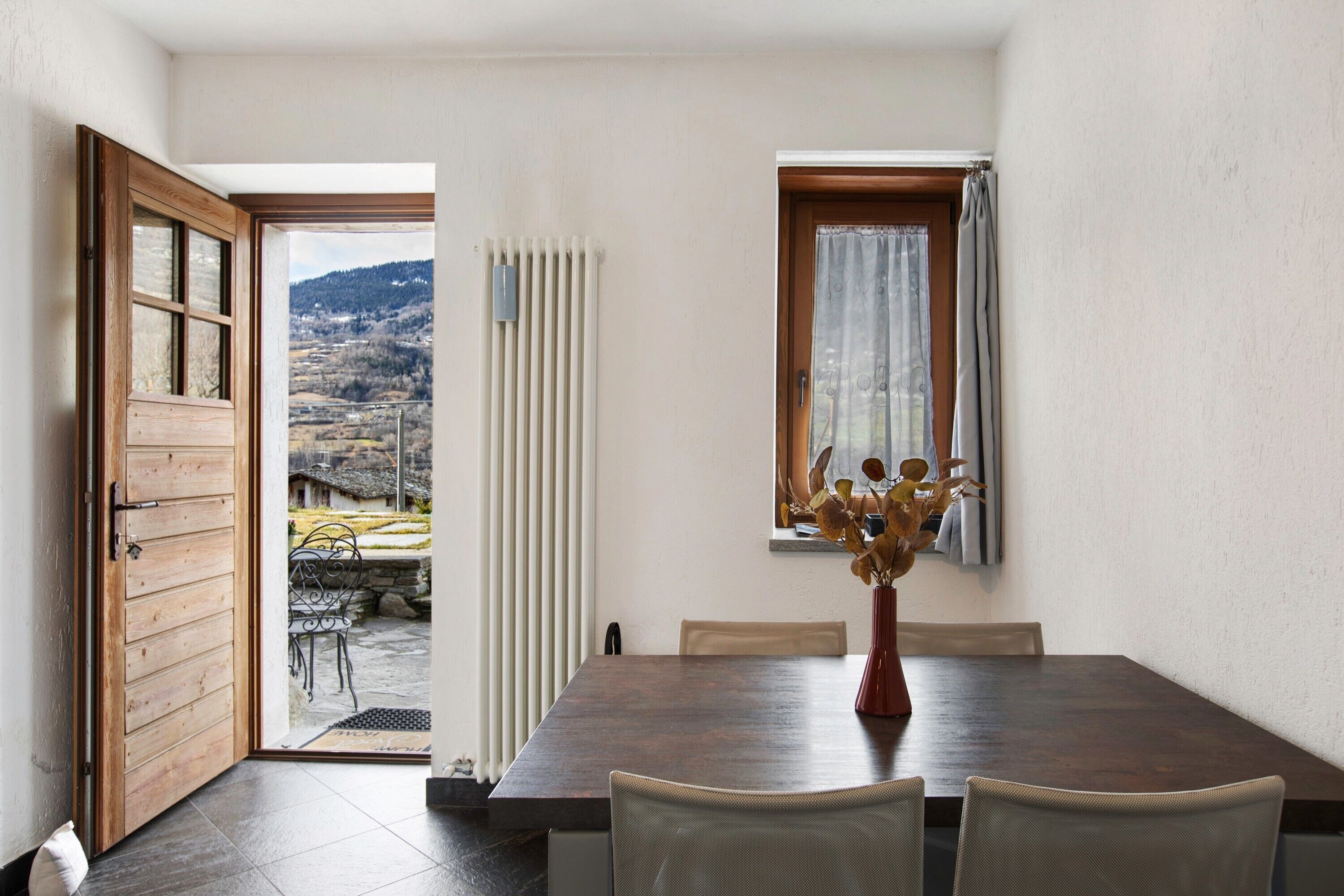 Un coin repas avec une table en bois, des chaises, un vase de fleurs séchées et une vue sur un paysage rural par une porte ouverte.