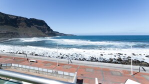 Vista costera con playa rocosa, cielo despejado y un edificio con tejado de tejas rojas.