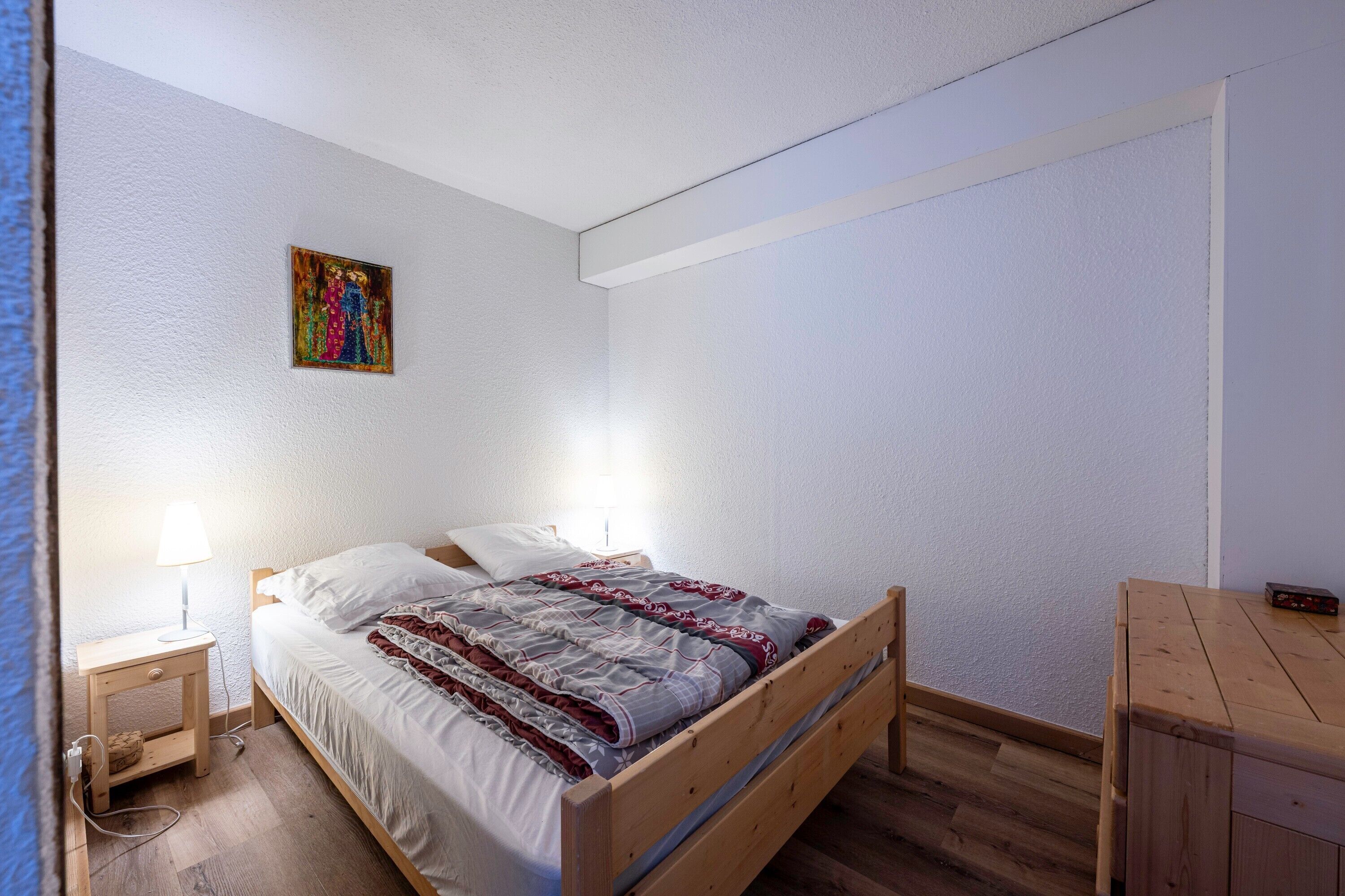 Een slaapkamer met een houten bed, een nachtkastje met een lamp, een aan de muur hangend schilderij en een klein houten tafeltje.