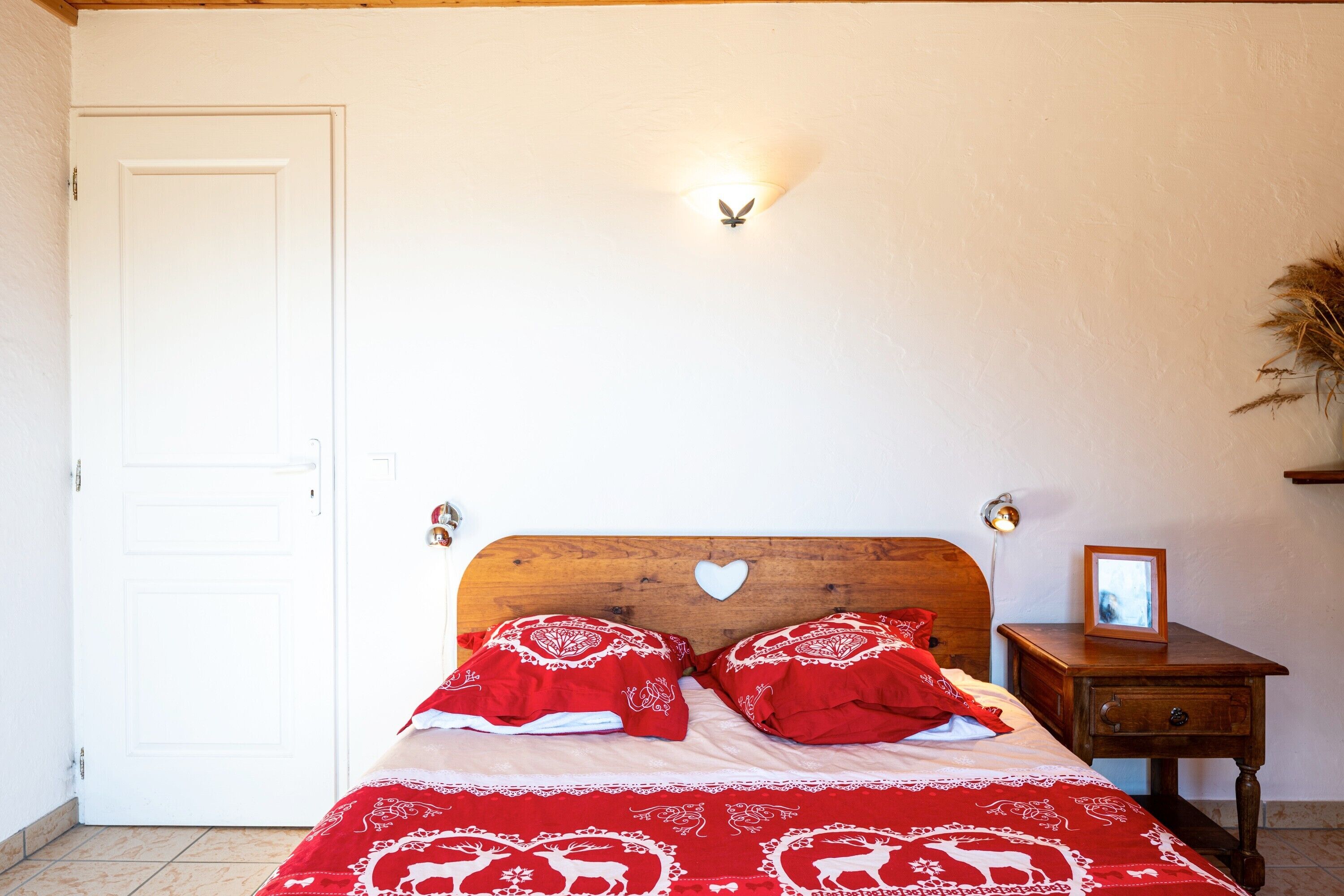 Een slaapkamer met een houten bed, rode beddengoed, een witte deur en een nachtkastje met een fotolijst.