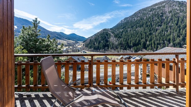 Un balcon doté d’une balustrade en bois, une seule chaise marron et une vue sur une vallée avec des montagnes et des maisons.