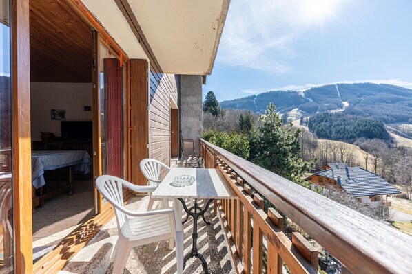 A balcony with a table and chairs, a wooden railing, and a view of a valley with houses and trees.