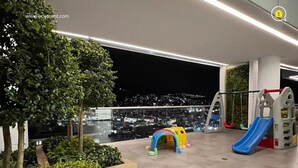 A rooftop terrace with a children's play area, including a slide and a climbing structure, surrounded by greenery and overlooking a cityscape at night. - 3 Dormitorios en el Edificio ONE Quito (Quito)
