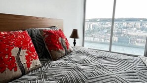 A bed with patterned bedding and red and black pillows, a bedside lamp, and a large window with a city view. - 3 Dormitorios en el Edificio ONE Quito (Quito)