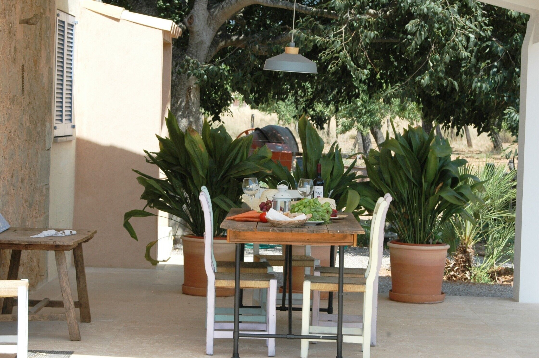 Un espacio de comedor al aire libre con una mesa lista para dos personas, rodeado de plantas en macetas y un colgante luminoso.