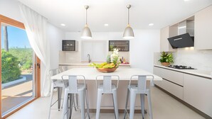 A modern kitchen with a central island, bar stools, pendant lighting, and a view of greenery outside. - Villa 'Can Americano, 4 with Mountain Views, Wi-Fi and Air Conditioning (Illes Balears)