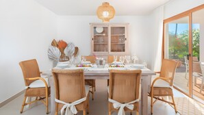 A dining area with a table set for six, surrounded by wicker chairs with white cushions, a white cabinet with glass doors, and a large window offering a view of greenery. - Villa 'Can Americano, 4 with Mountain Views, Wi-Fi and Air Conditioning (Illes Balears)