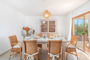 Un coin repas avec une table dressée pour six personnes, entouré de chaises en osier avec des coussins blancs, d’un buffet blanc aux portes en verre et d’une grande fenêtre offrant une vue sur la verdure.