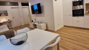 Una cucina moderna con mobili bianchi, un forno a microonde e un forno. C'è una zona pranzo con un tavolo e sedie bianche, e una TV montata a parete.