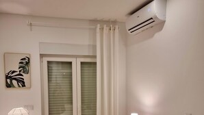 Camera da letto con un letto, cuscini, una lampada da comodino, un condizionatore d'aria, una finestra con tende e un'opera d'arte appesa al muro.