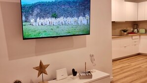 Un soggiorno moderno con TV a schermo piatto montata a parete, un mobile basso bianco e un angolo cottura con pensili bianchi e un piano di lavoro in legno.