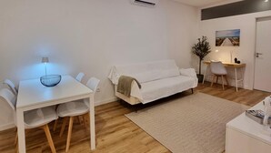 Un soggiorno moderno con un divano bianco, un tavolo da pranzo con sedie, una piccola scrivania con sedia e un condizionatore d'aria a soffitto.