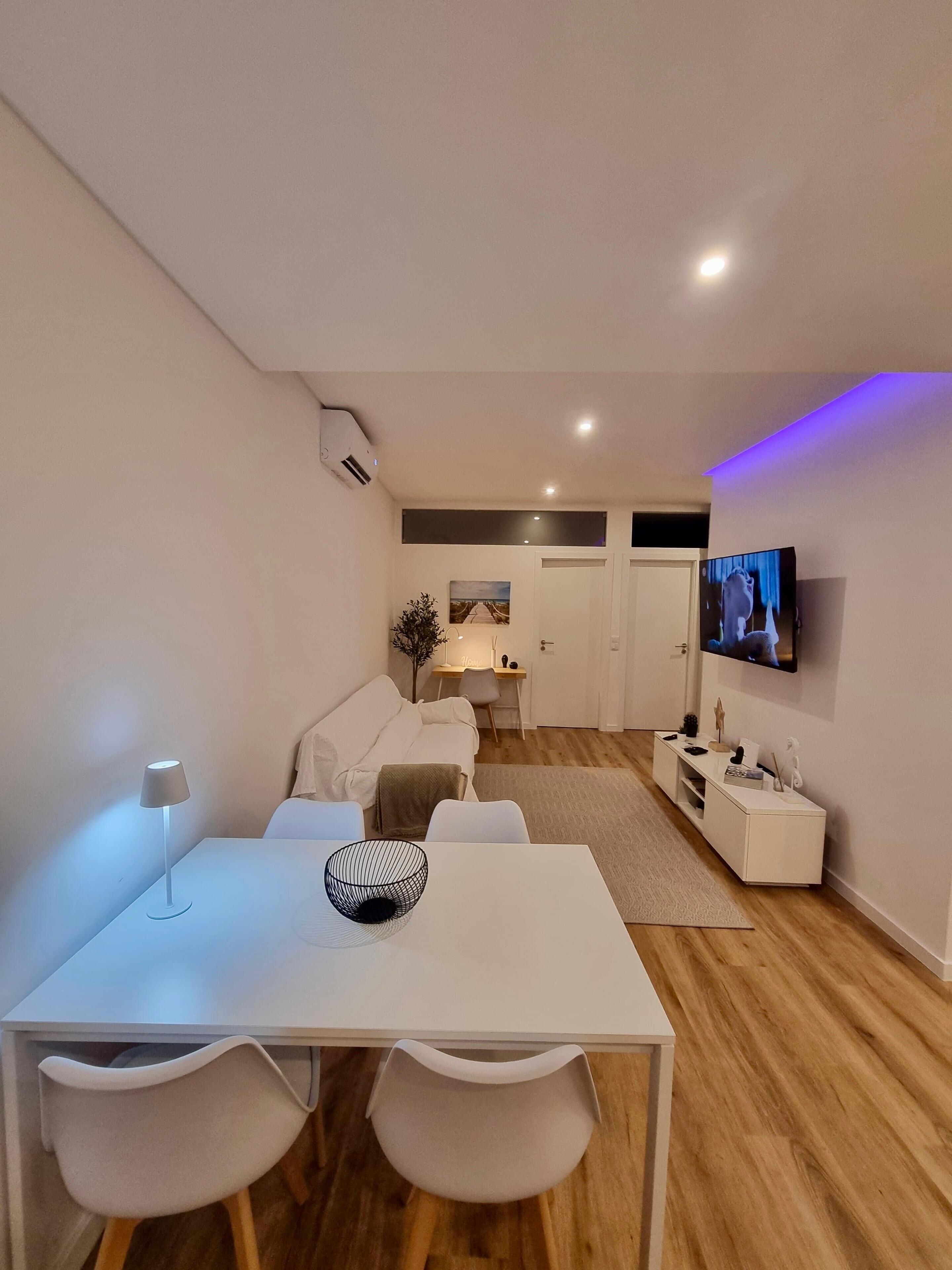 A modern, well-lit living room with a white sofa, a dining table with two chairs, a TV mounted on the wall, and a small white side table with decorative items.