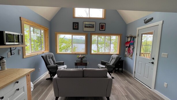 Una habitación con techo estilo loft, dos ventanas con vista a un lago, un sofá gris y una mesa de madera.