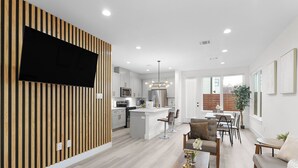 A modern kitchen with a central island, white cabinetry, and a flat-screen TV mounted on a wooden slat wall. - Modern Heights Townhome 3BR with Fast Wifi & Free Park (Houston)