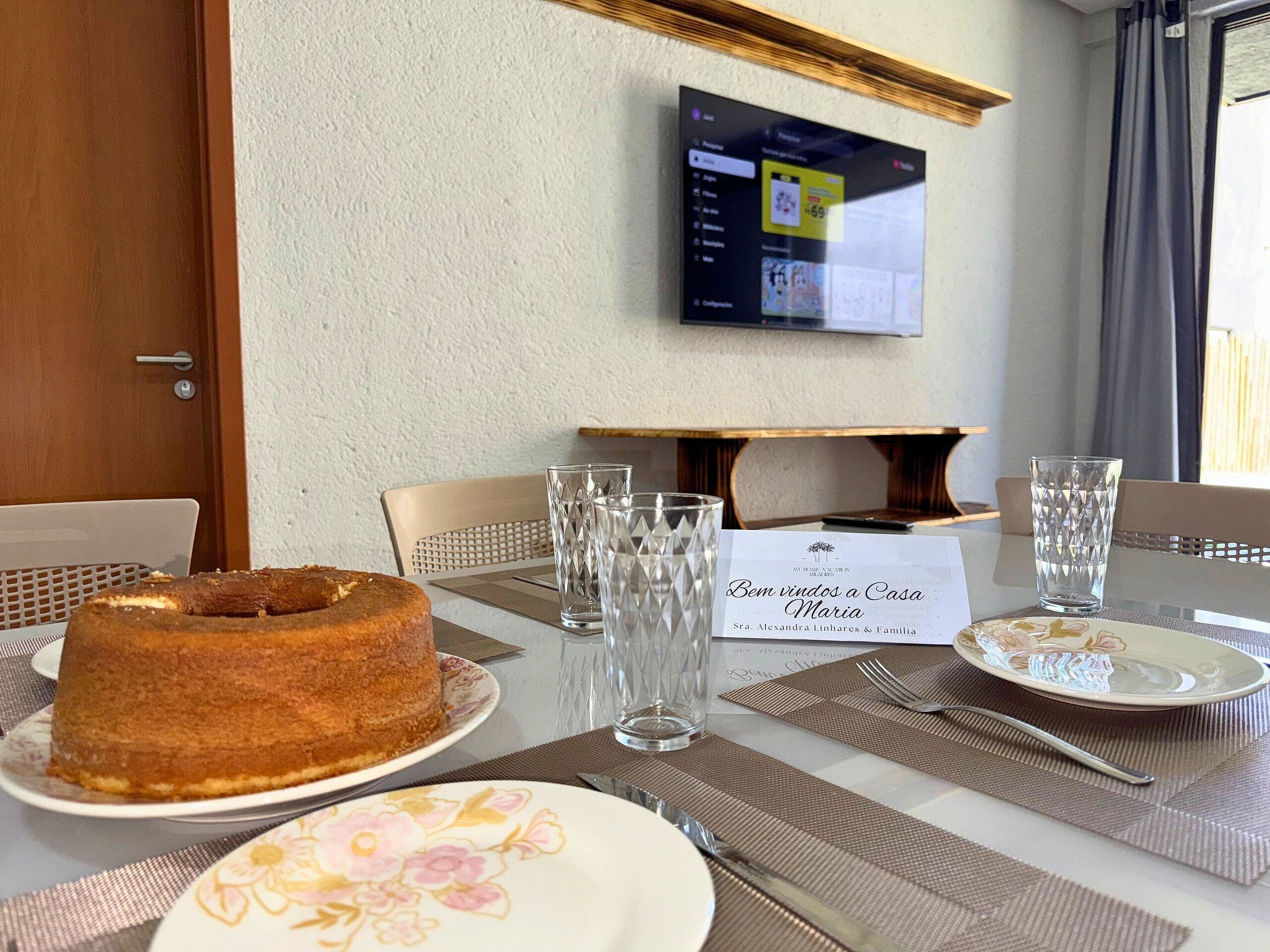 A dining table with a cake, glasses, and a menu, with a TV mounted on the wall in the background.