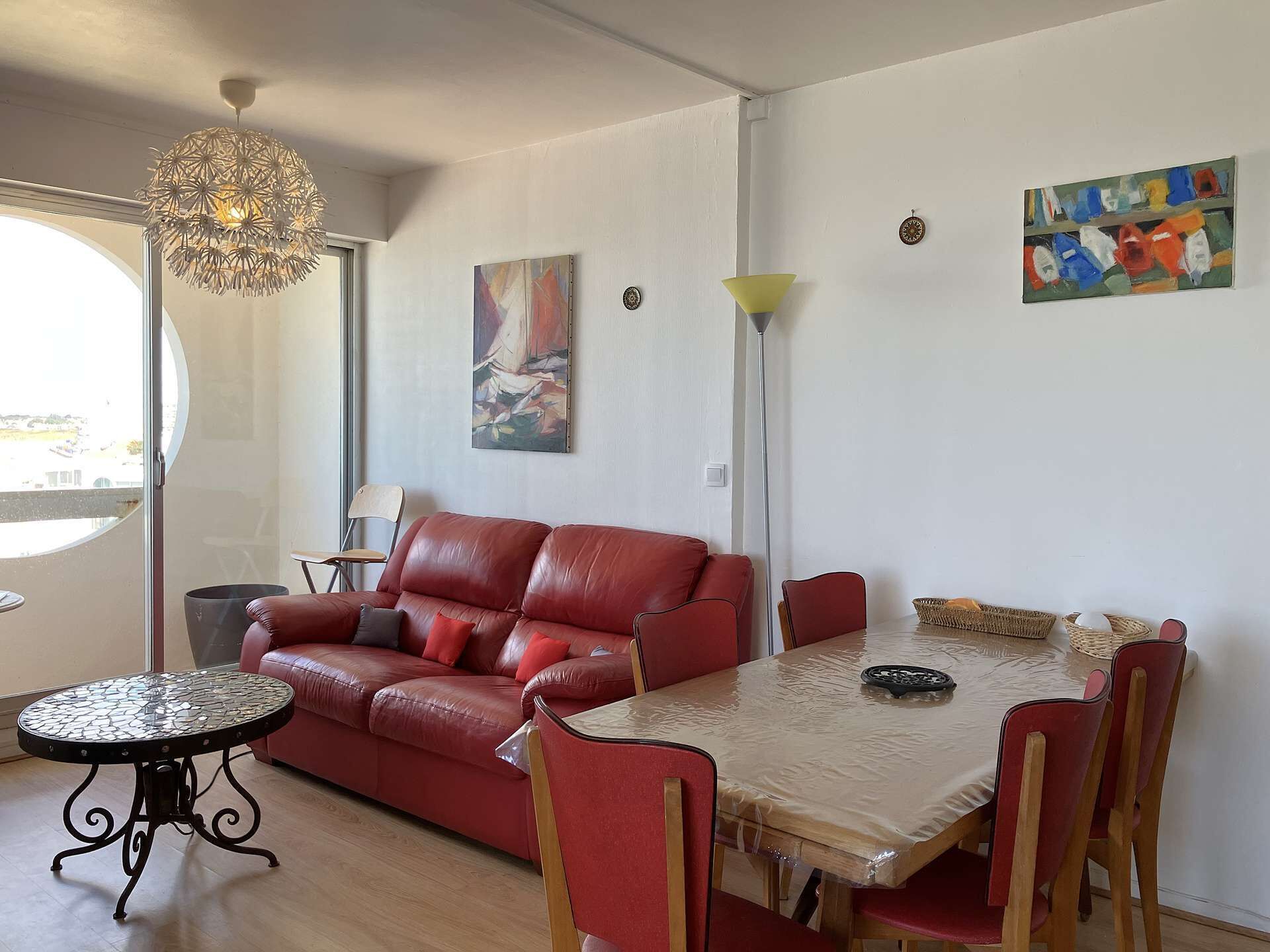 Un salon avec un canapé en cuir rouge, une table ronde et une table à manger avec des chaises. Il y a un tableau au mur et un lampadaire.