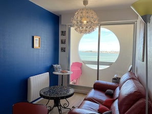 Una habitación con pared azul, una mesa redonda con revistas, un sofá rojo, una lámpara colgante y vista al mar a través de un ventanal circular.