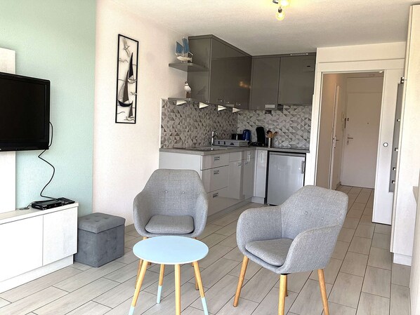 A compact living space with a kitchenette, a dining area with two grey chairs and a small round table, and a flat-screen TV mounted on the wall.