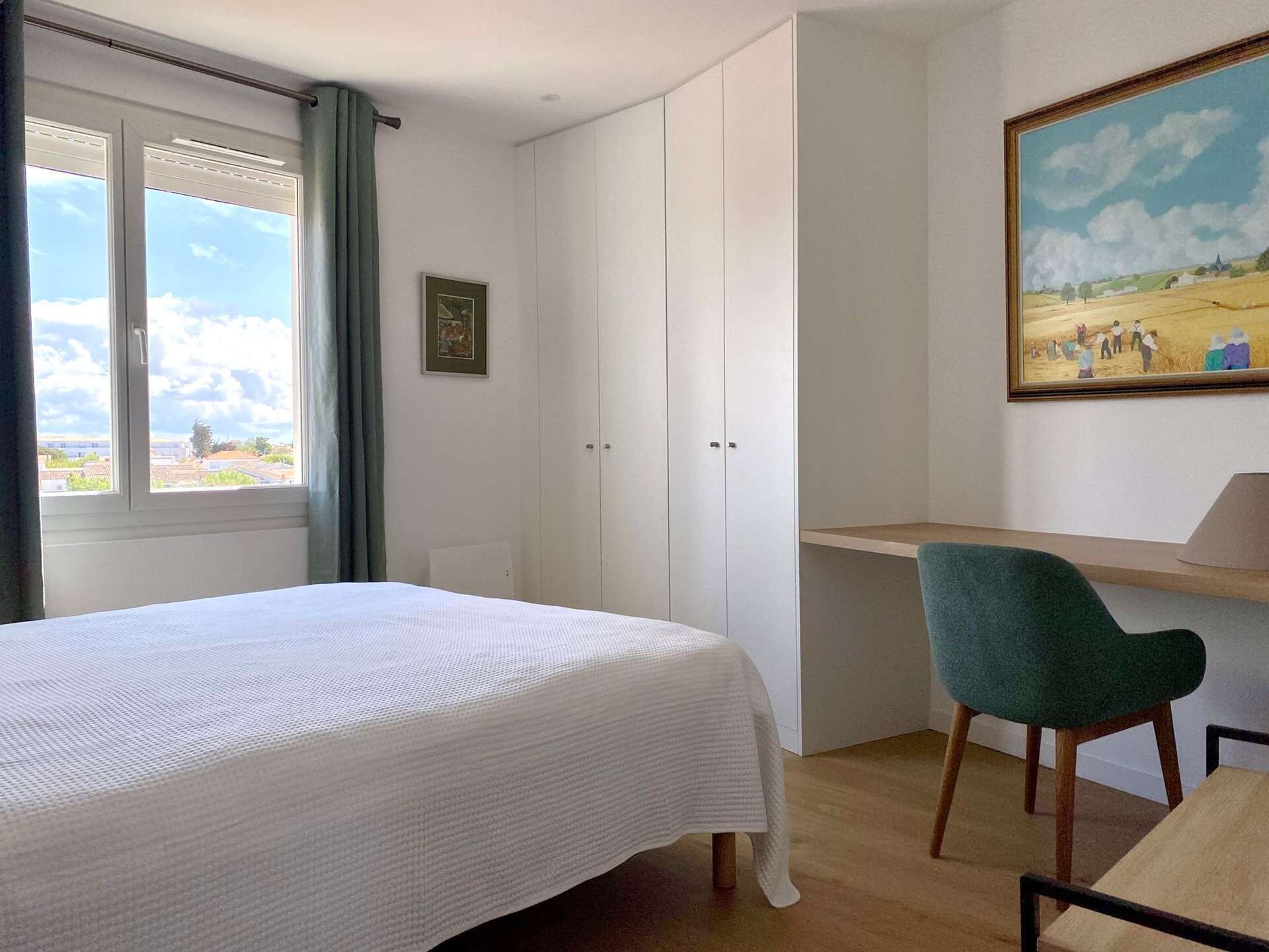 A bedroom with a bed, a desk, a chair, a painting, and a window with a view of the sky and clouds.
