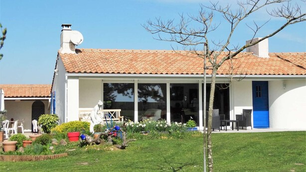 A white building with a tiled roof, a blue door, and a covered patio area with outdoor furniture. - Les Treilles Saint Hilaire de Riez (SAINT HILAIRE DE RIEZ)