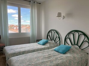 Et soveværelse med to enkeltsenge, et vindue med udsigt til bygninger, en radiator og en vægmonteret lampe.