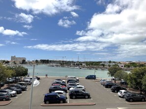 Marina com carros estacionados e barcos, céu claro e edifícios ao fundo.