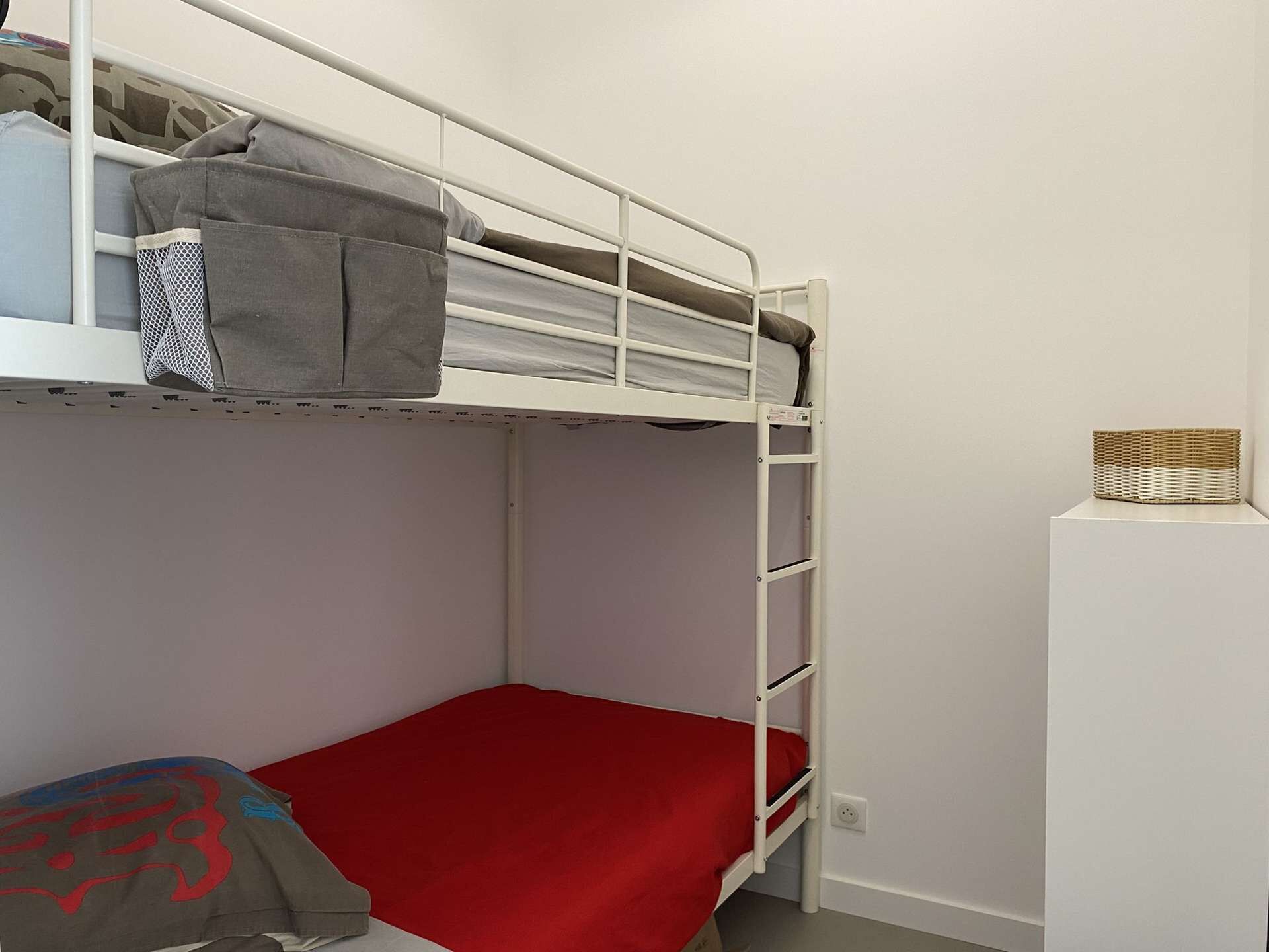 Una cama individual con colchón rojo, estructura metálica blanca y un pequeño estante blanco con una cesta de mimbre.