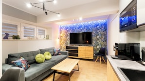 A modern living room with a sofa, coffee table, and a TV. There's a kitchen area with a microwave and a coffee machine.