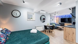 A modern hotel room with a large bed, a dining area, a TV, and a kitchen.