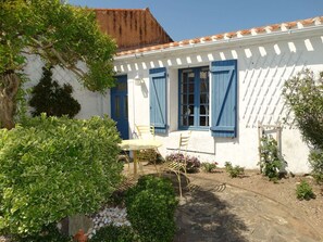 A white building with blue shutters and a tiled roof. There is a table and chairs outside, surrounded by greenery.