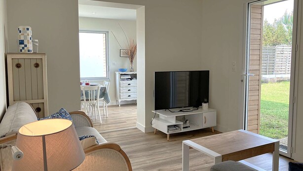 Un salon moderne avec un téléviseur, un canapé et une lampe. On y trouve également un coin repas avec des chaises et une table, et une vue sur un jardin grâce à une porte coulissante.