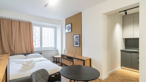 A compact hotel room with a bed, a small desk, a chair, a round table, and a kitchenette.