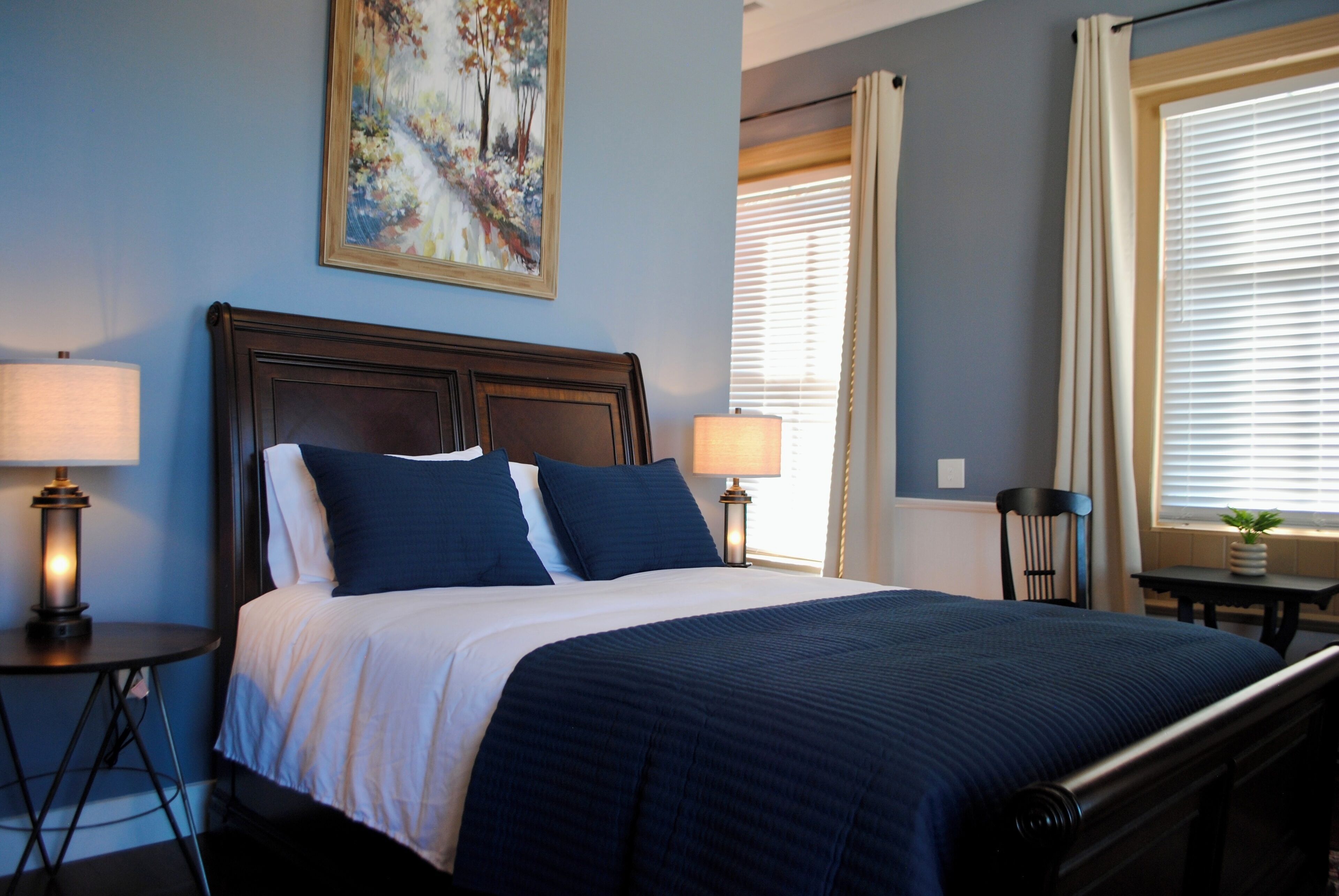 Una cama bien hecha con cubrecama y almohadas azul oscuro, cabecera de madera, dos lámparas de noche, un cuadro en la pared y una ventana con persianas.