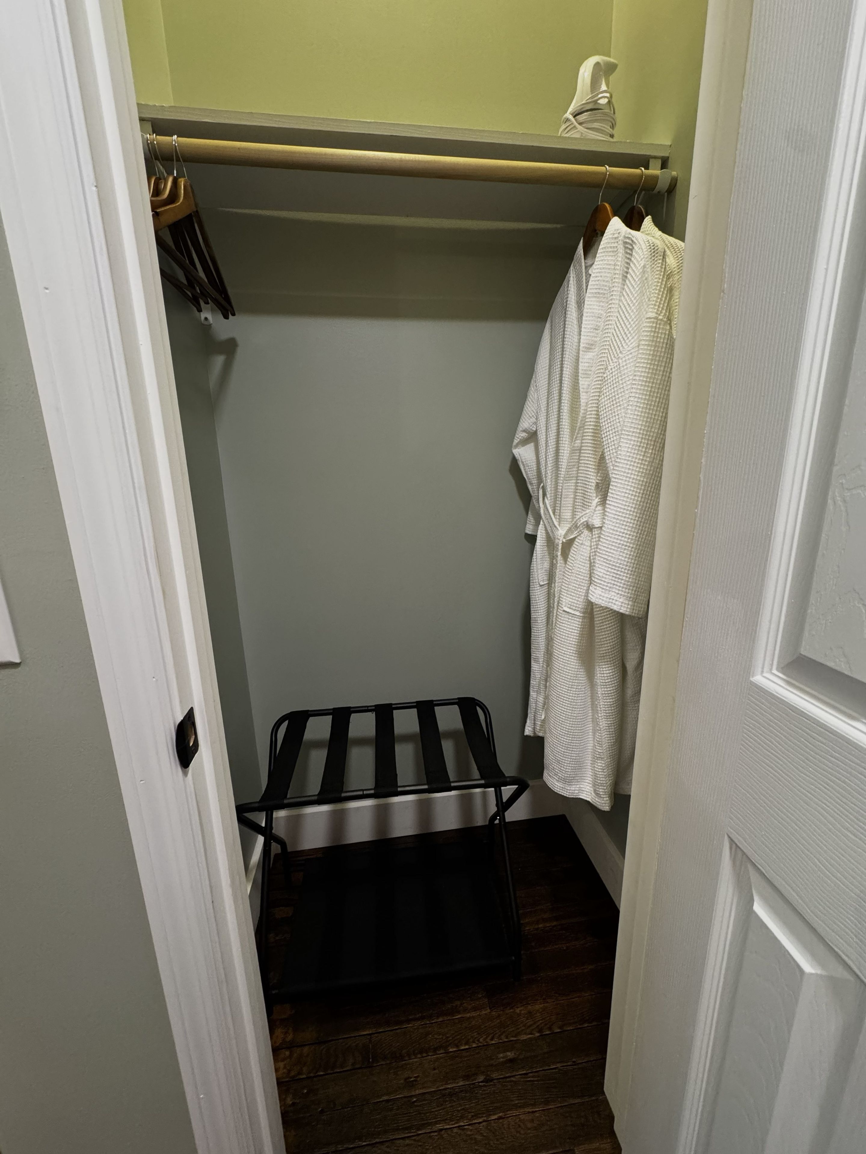 A narrow hallway with a wooden bench, a clothing rack with a white robe, and a door on the left.