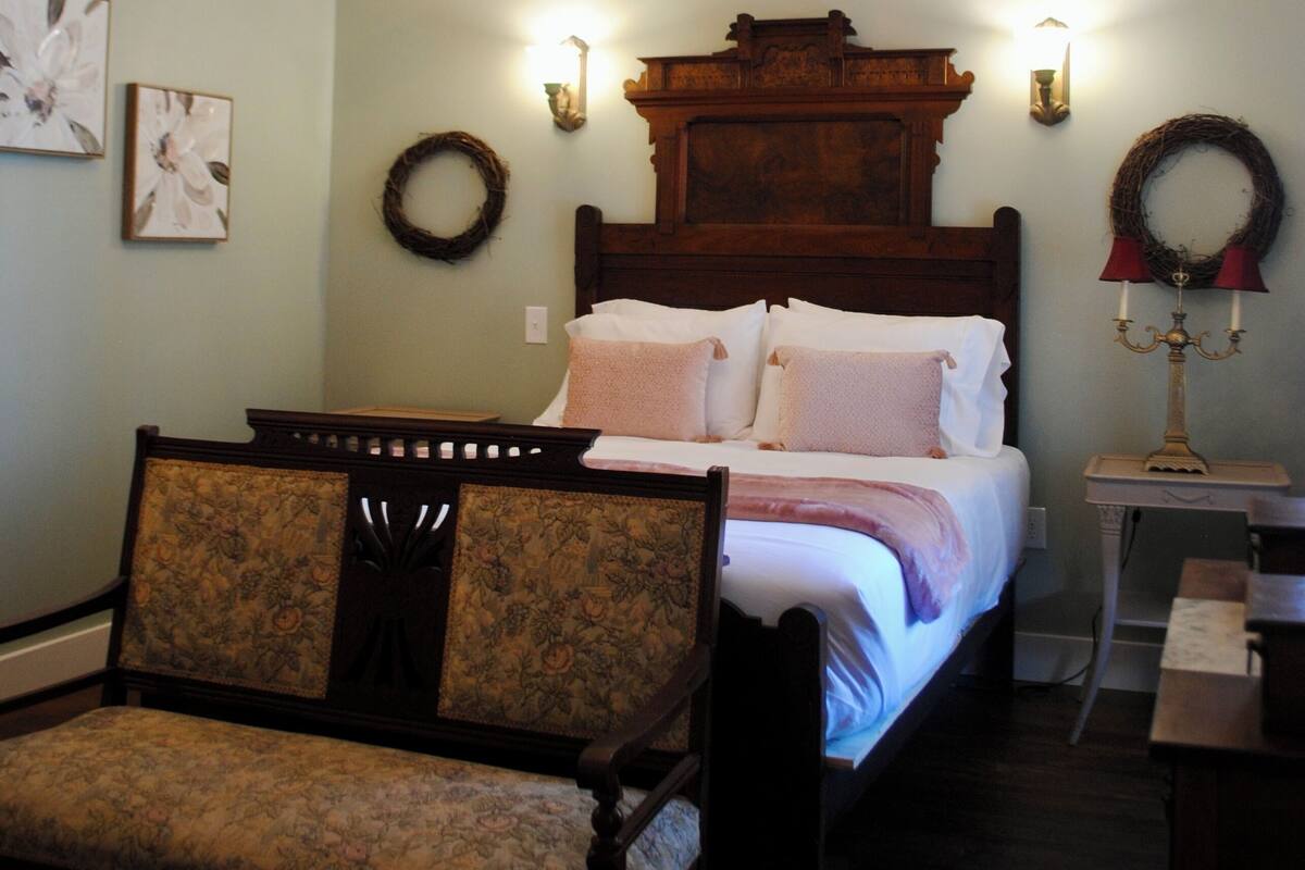 Ein Schlafzimmer mit einem hölzernen Bett, einem Sessel, einer Chandelierleuchtre und Wandleuchten sowie einem kleinen Tisch.