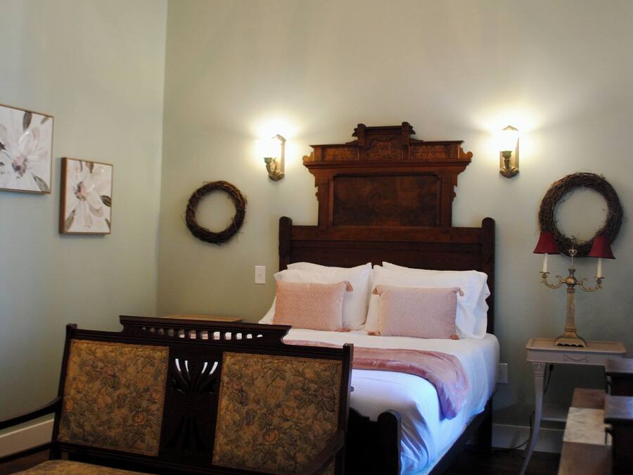 Un dormitorio con cama de madera, una silla, una araña y apliques de pared, junto a una mesita.