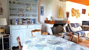 A cozy living room with a round table covered in a blue and white patterned tablecloth, a white cabinet with decorative plates, a fireplace with a small sailboat decoration, and a television on a wooden stand. - Frégate Saint Hilaire de Riez (SAINT HILAIRE DE RIEZ)