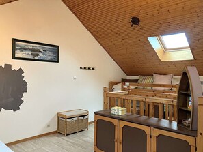 Une chambre à coucher aménagée dans un espace de type « loft », avec un plafond en bois, un lit superposé, un puits de lumière, une grande œuvre d’art abstraite murale et un petit meuble en bois.