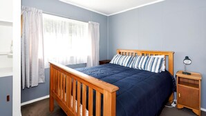 A bedroom with a wooden bed, a nightstand, a lamp, and a window with curtains. - Okiwi Bay Bach with Bay Views (Ōkiwi Bay)