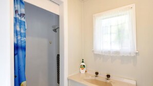 A bathroom with a shower, a sink with a mirror, and a window with a white curtain. - Okiwi Bay Bach with Bay Views (Ōkiwi Bay)