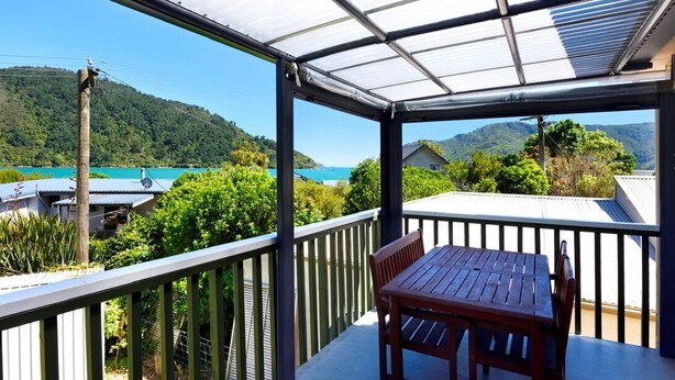 A covered outdoor area with a table and chairs, overlooking a scenic view of a lake and hills. - Okiwi Bay Bach with Bay Views (Ōkiwi Bay)