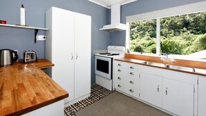 A modern kitchen with white cabinets, a wooden countertop, a stove, and a window with a view of greenery. - Okiwi Bay Bach with Bay Views (Ōkiwi Bay)