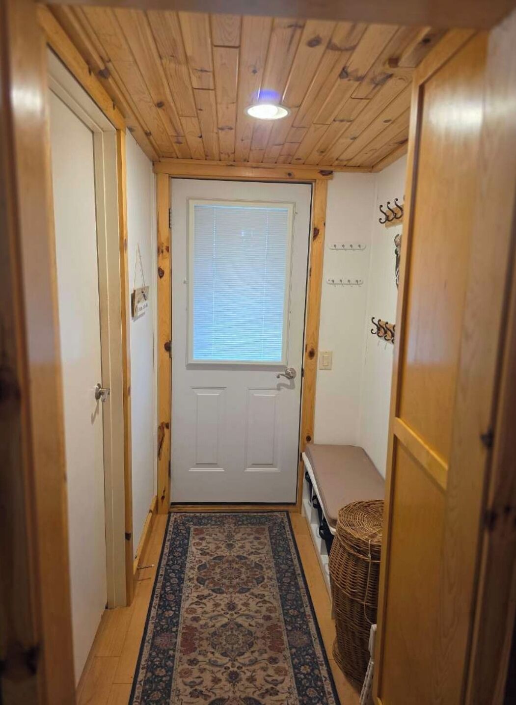 A narrow hallway with wooden walls and a door with a window. A patterned rug runs along the floor, and a wicker basket is placed against the wall.