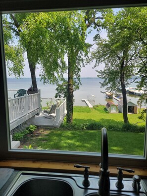 A view from a window overlooking a grassy area with a dock and boats, a wooden deck with chairs, and a small shed. - Cozy 3-bedroom cabin in wonderful Perham with WiFi, AC and great fishing! (Perham)