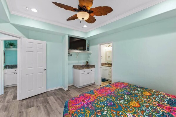 A bedroom with a bed, a ceiling fan, a TV, and a bathroom visible through an open door. - Dragonfly & Flamingo (Gilchrist)