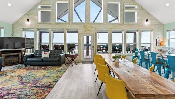 A spacious loft-style room with a high ceiling, large windows, a wooden dining table, yellow chairs, and a cozy seating area with a sofa and a TV.