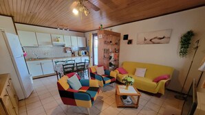 A living room with a yellow sofa, colorful armchairs, and a coffee table. There is a kitchen in the background with white cabinets and a dining table.