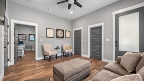 A spacious living room with a ceiling fan, a large ottoman, and two armchairs. There are framed certificates on the wall, and a door leads to another room.